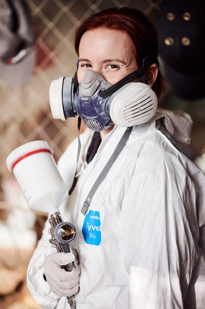 Eine Frau hat eine Atemschutzmaske aufgesetzt und hält eine Lackierpistole.